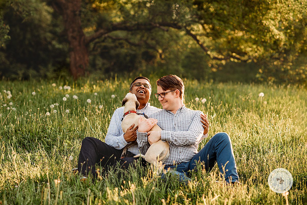 two men laugh with their dog licking them in field during Jamaica Plain family portraits