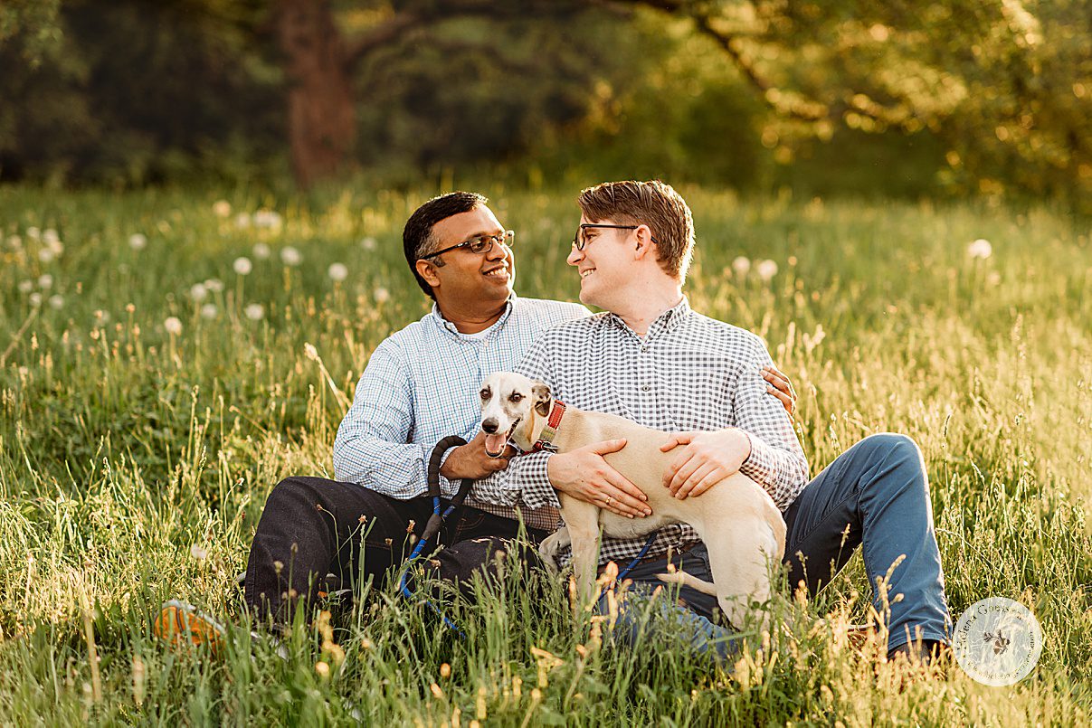 husbands smile together in tall grass field during Jamaica Plain family portraits while holding dog 
