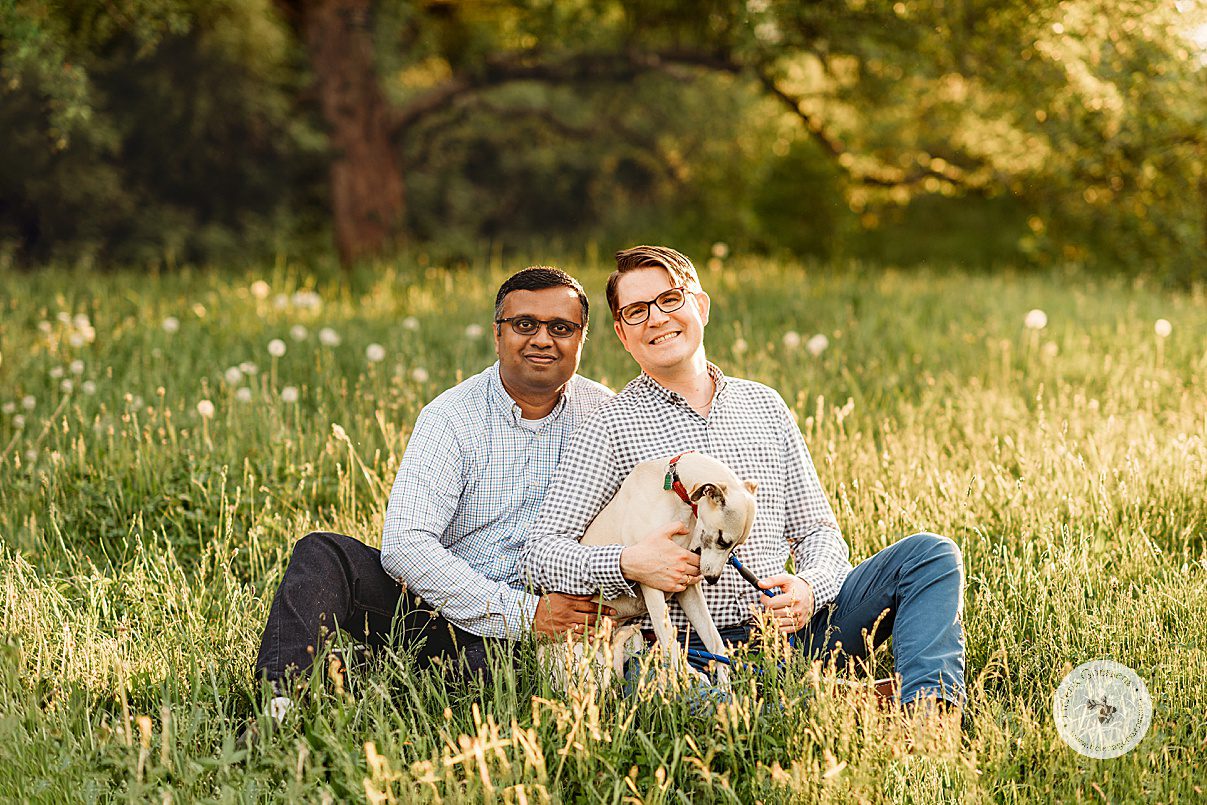 married couple sits in grass with dog licking their hands during Jamaica Plain family portraits