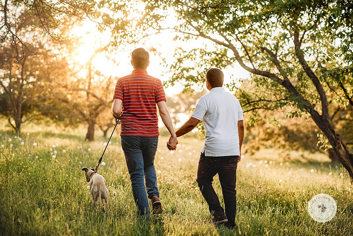 husbands hold hands walking up hill during Jamaica Plain family portraits with dog on leash next to them 