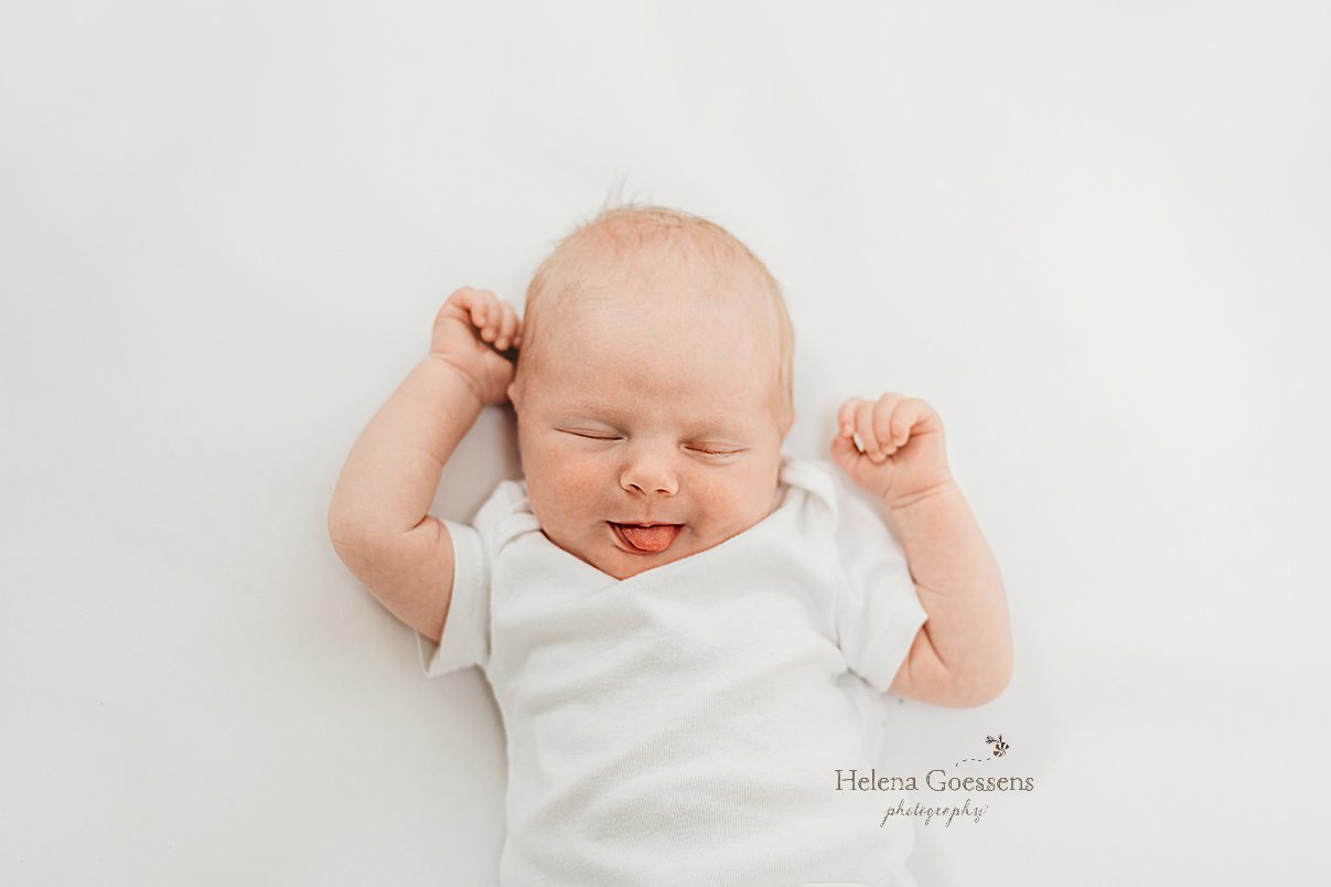 baby girl stretches in white onesie sticking her tongue out during lifestyle newborn portraits in Dedham MA