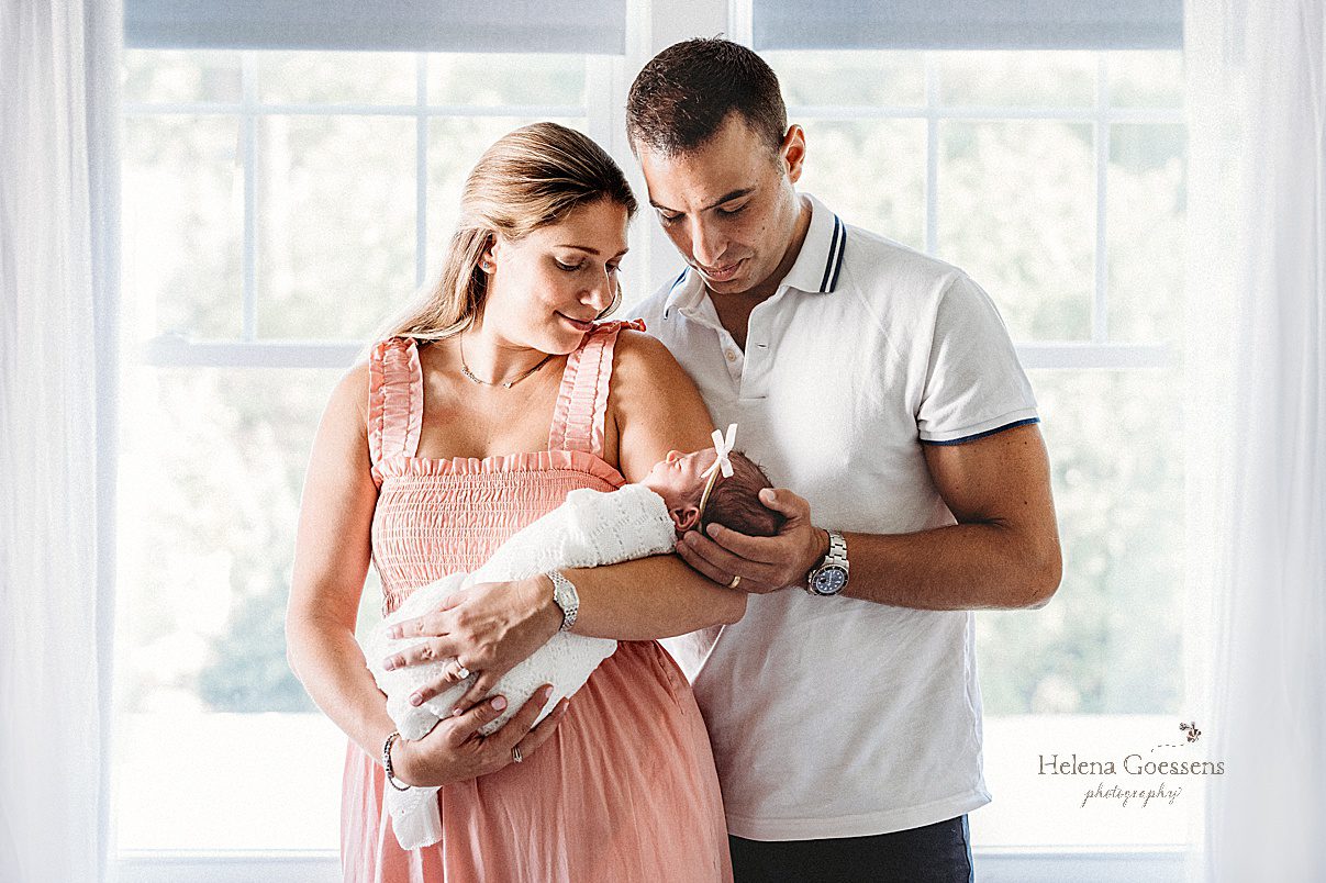 parents look down at baby girl standing by windows during Newborn Session in Needham MA