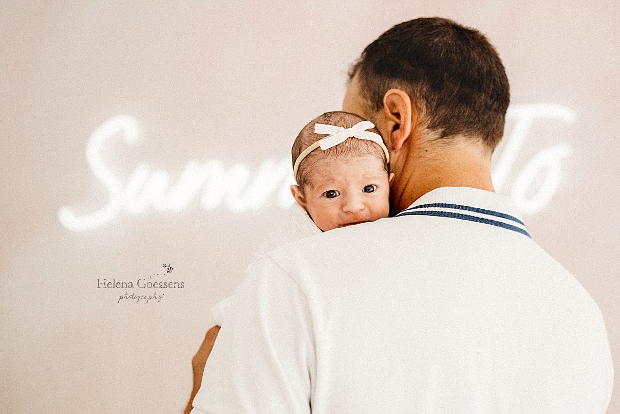 dad holds newborn baby girl laying on his shoulder in front of neon sign during MA newborn photos 