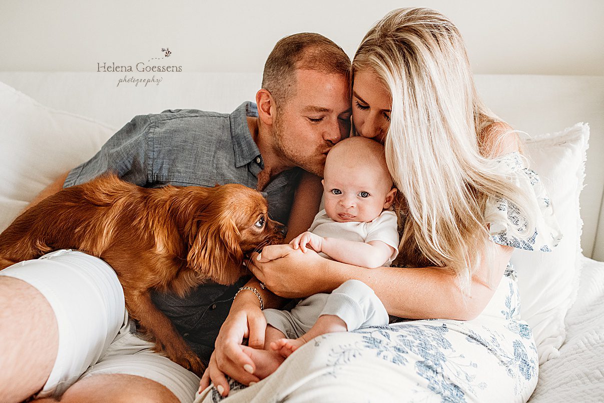 parents kiss baby boy while snuggling with dog in bed photographed by Helena Goessens Photography 