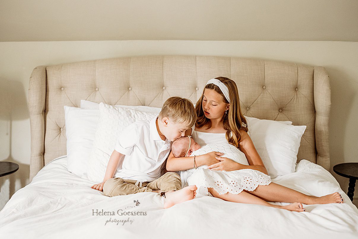 big sister holds baby while brother with Down syndrome kisses baby girl sitting on tan bed with white sheets