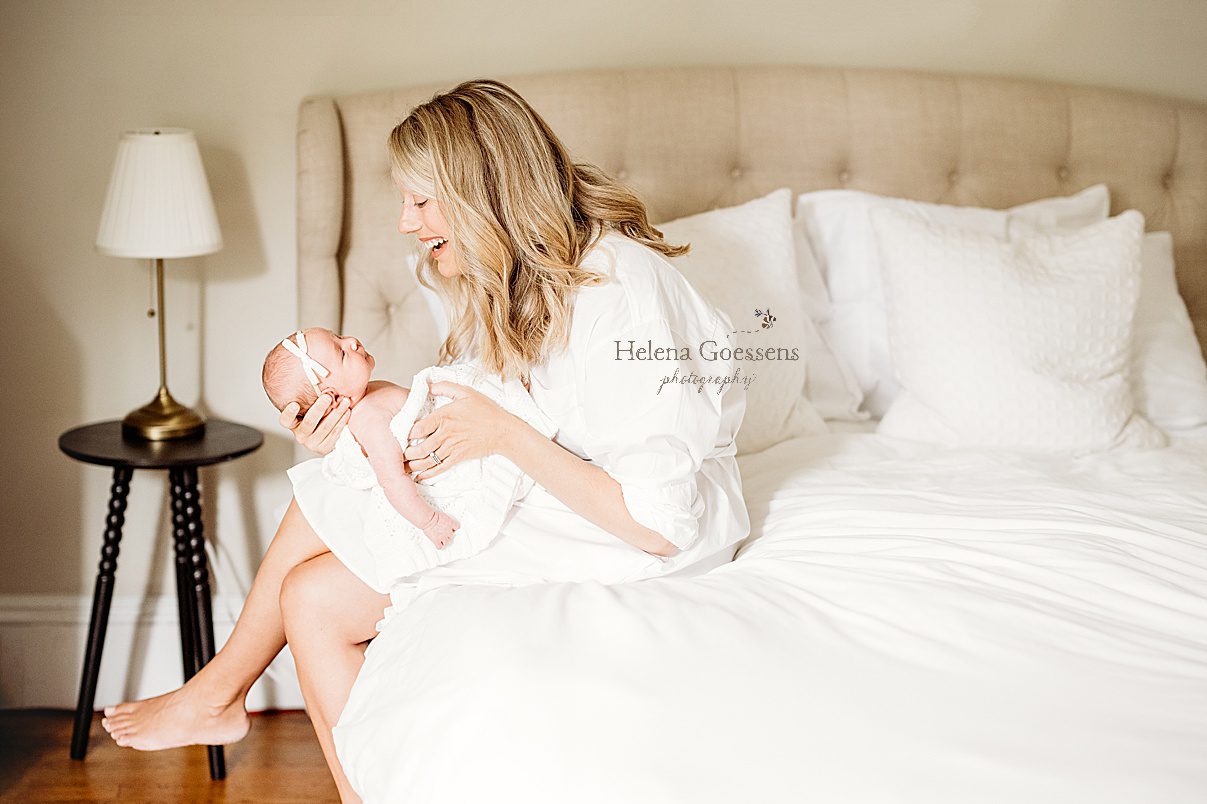mom laughs looking down at baby girl sitting on bed during lifestyle newborn session in Newton