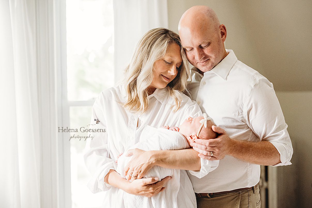 parents look down at baby girl with simple white bow on head during lifestyle newborn session in Newton