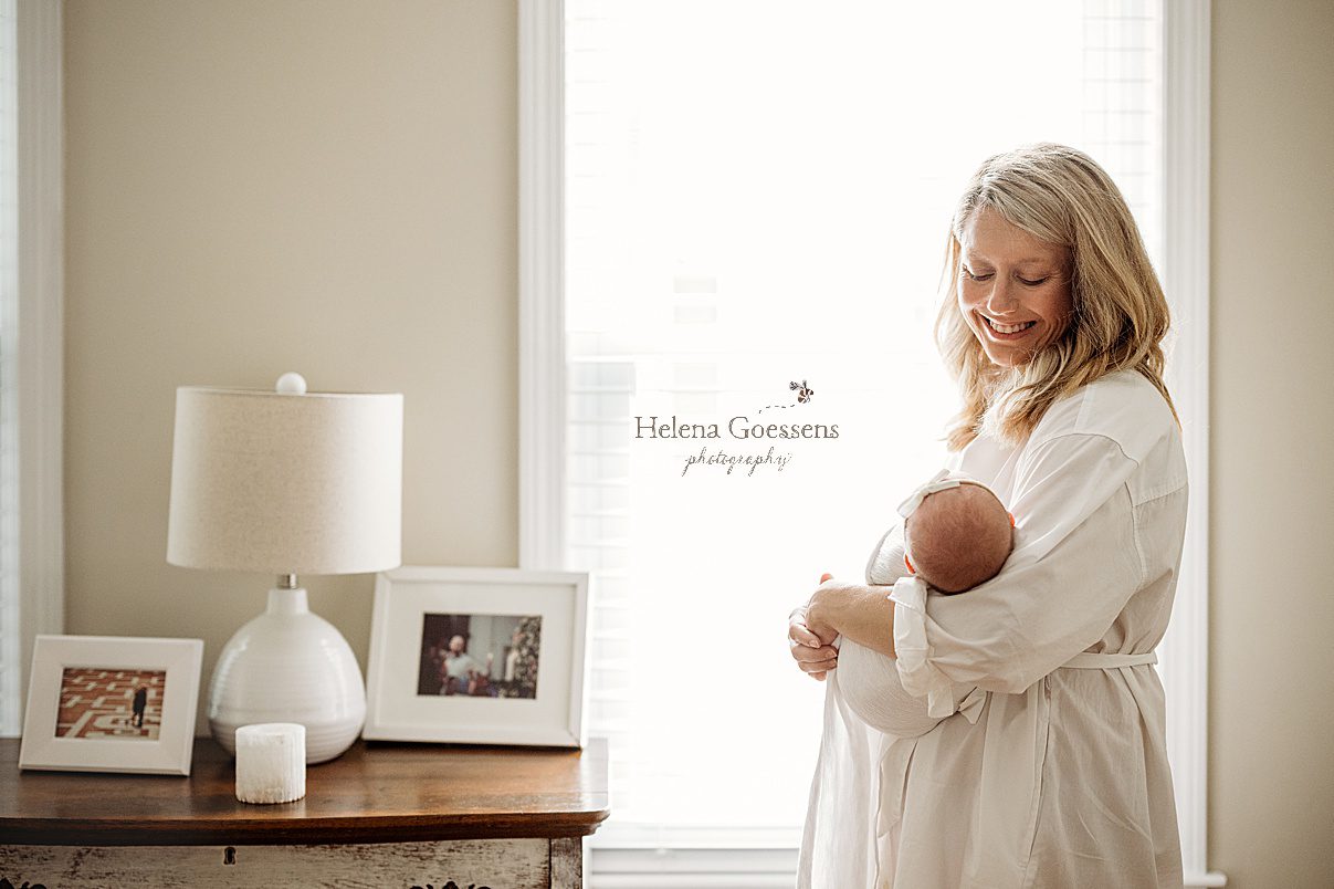 mom in white dress smiles down at baby girl in Newton MA home during lifestyle newborn session