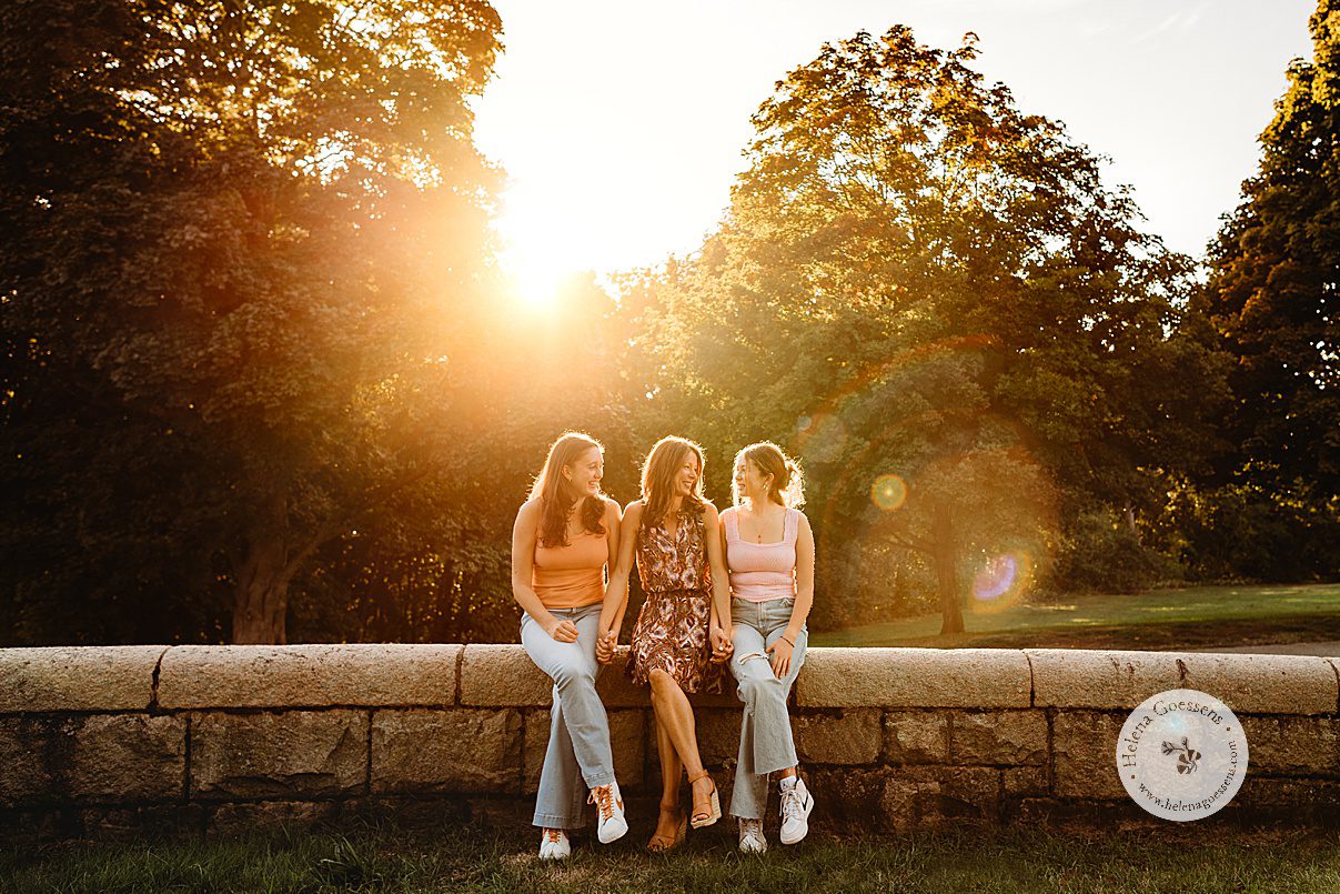 mom sits with two daughters on stone wall in Larz Anderson Park with sun flare through trees