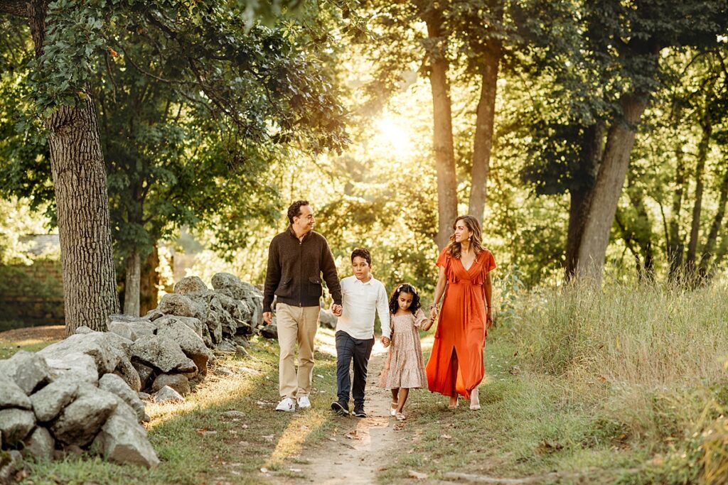 A Concord MA family portrait comes to life as a family of four, including two adults and two children, walks hand in hand along a path in the sunlit forest.