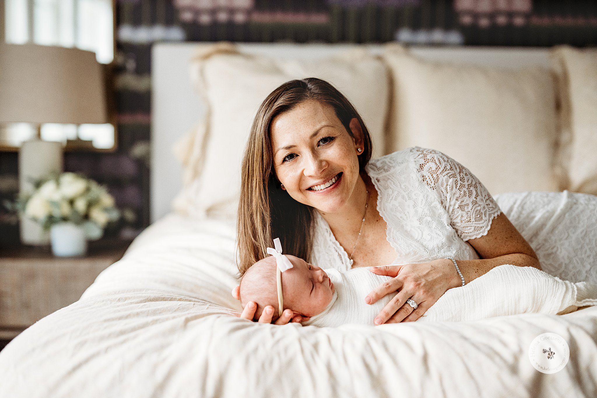 mom in white dress with lace sleeves lay on bed with baby girl in white wrap 