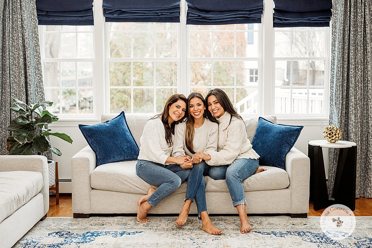 three sisters sit on tan couch with blue pillows in matching cream sweaters with jeans during generational family portraits in Boston MA