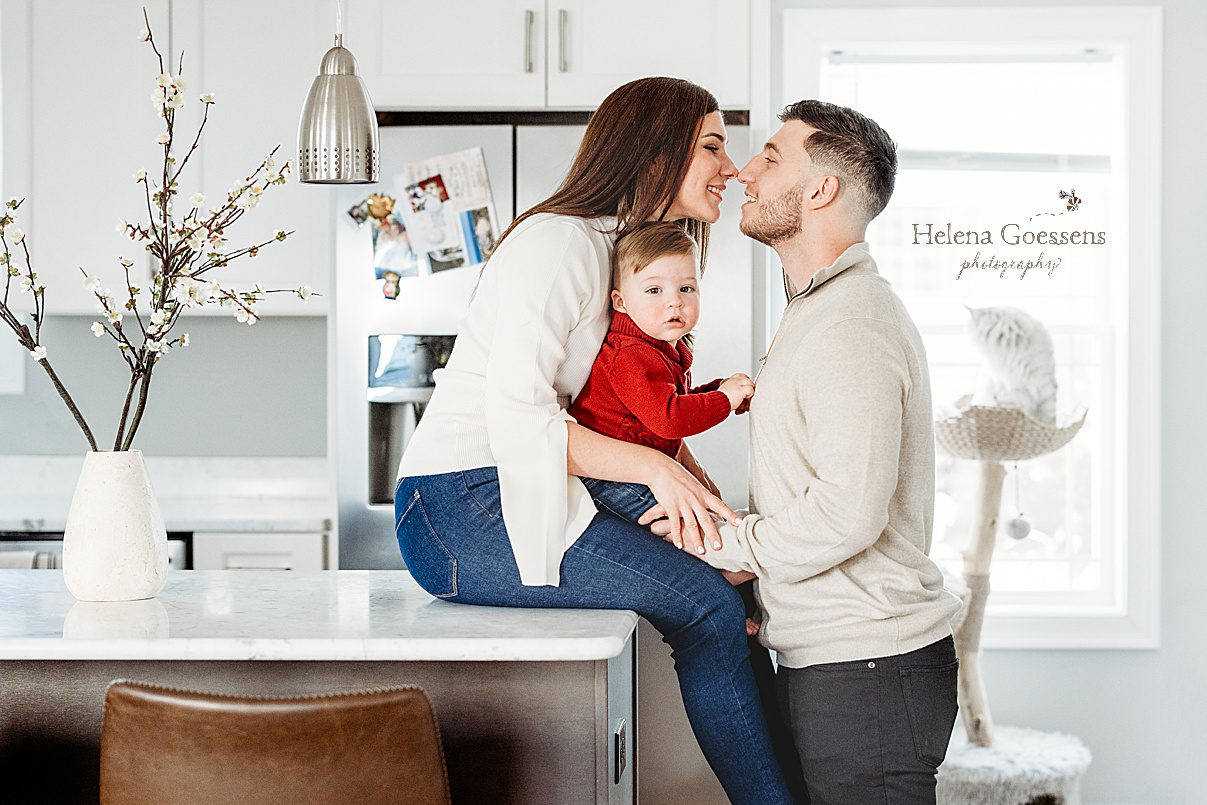 mom sits on counter with son on lap leaning to kiss husband during lifestyle family photos at home in Boston MA