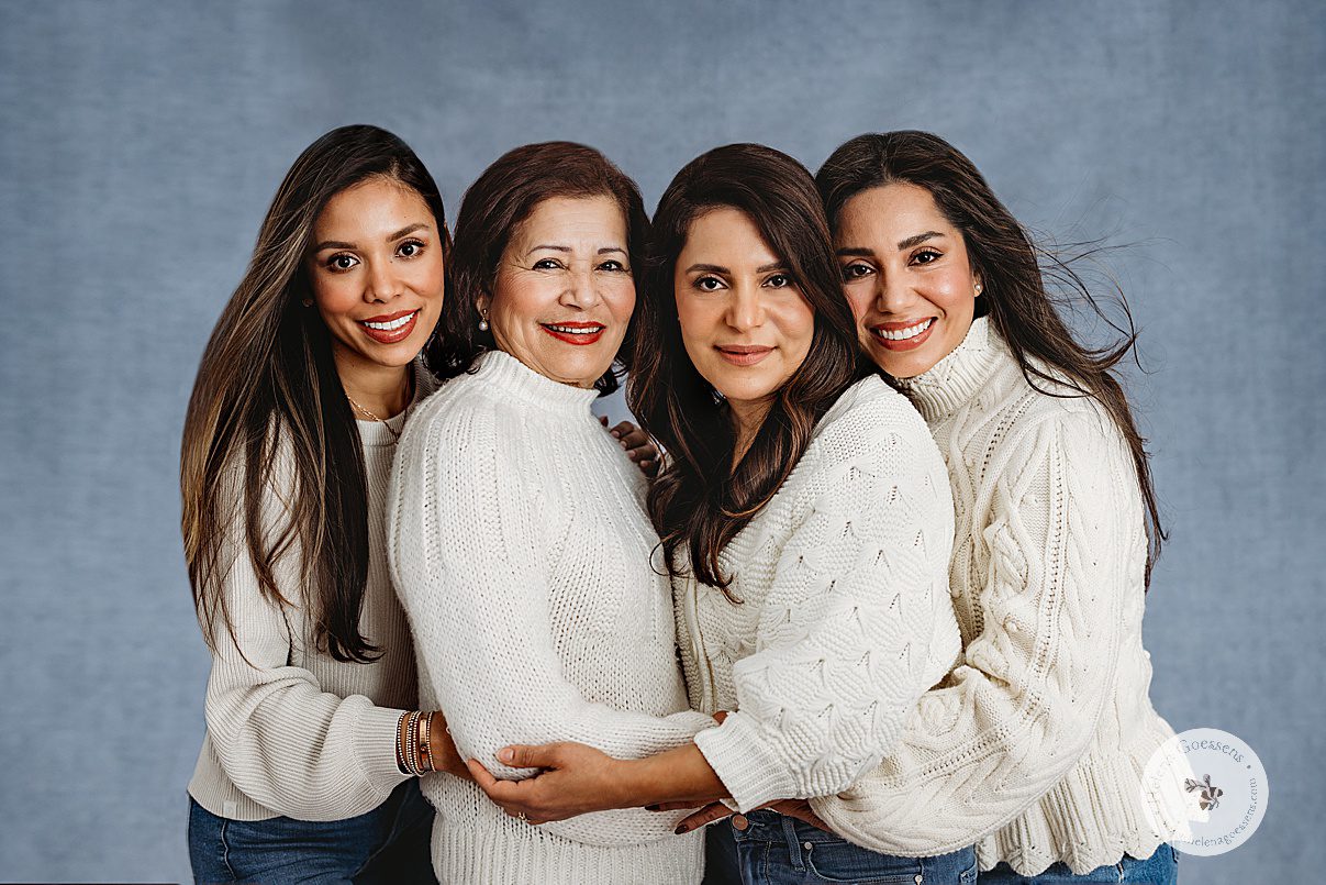 three sisters in cream sweaters and jeans hug mom in front of blue backdrop