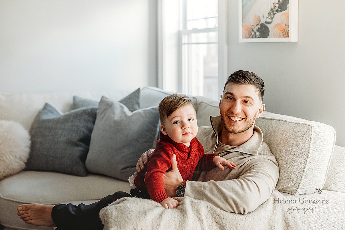 dad sits on couch with son in lap during lifestyle family photos at home in Boston MA