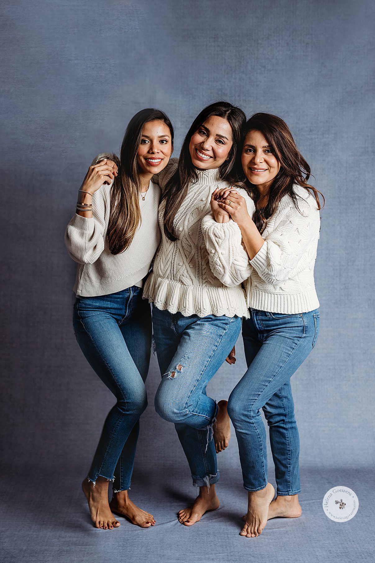three women in cream sweaters and jeans hug on blue backdrop during generational family portraits in Boston MA