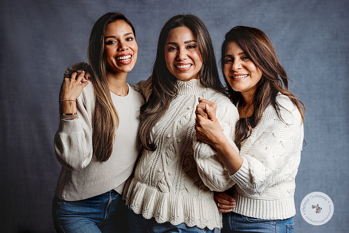three women hug and pose in cream sweaters on blue backdrop during generational family portraits in Boston MA