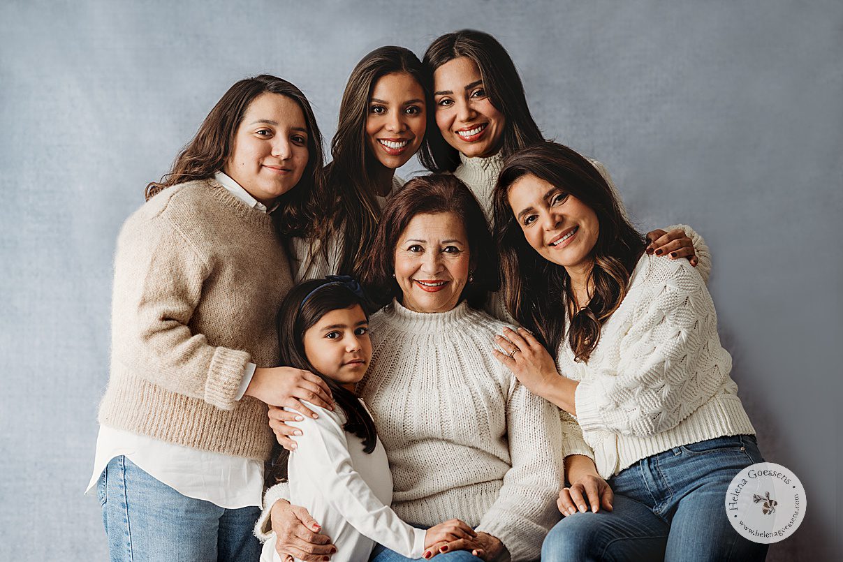 three women hug and pose in cream sweaters with mother and two daughters on blue backdrop during generational family portraits in Boston MA
