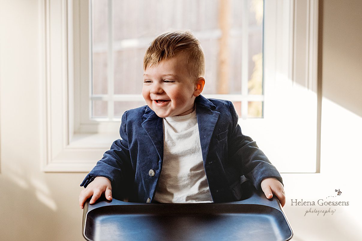 toddler sits in white shirt with blue jacket in high chair during first birthday photos at home 