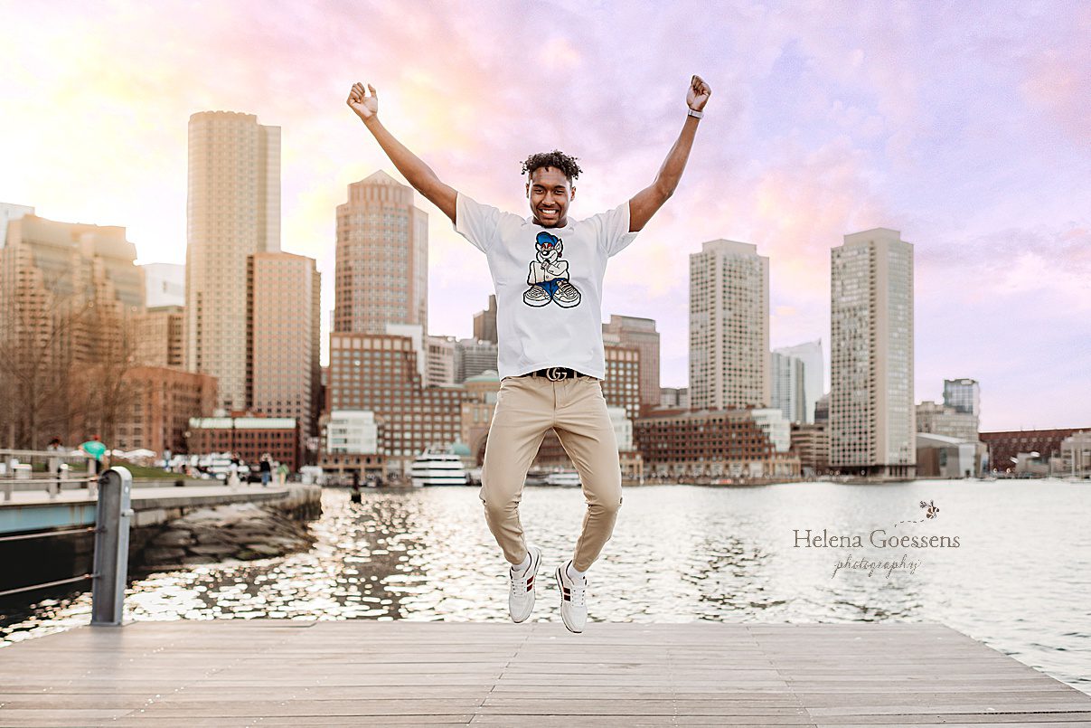 senior boy jumps on pier with Boston skyline behind him 