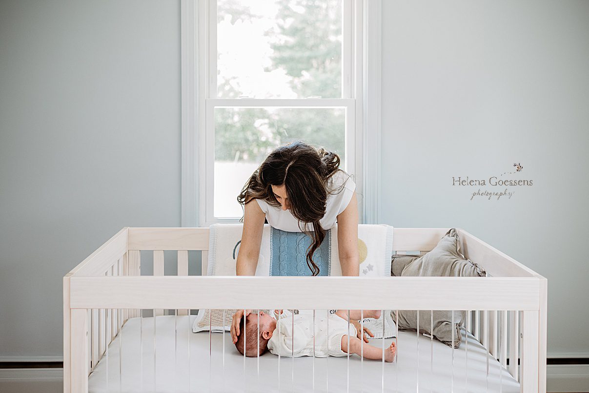 mom leans over side of white crib during newborn photos at home in Westwood MA