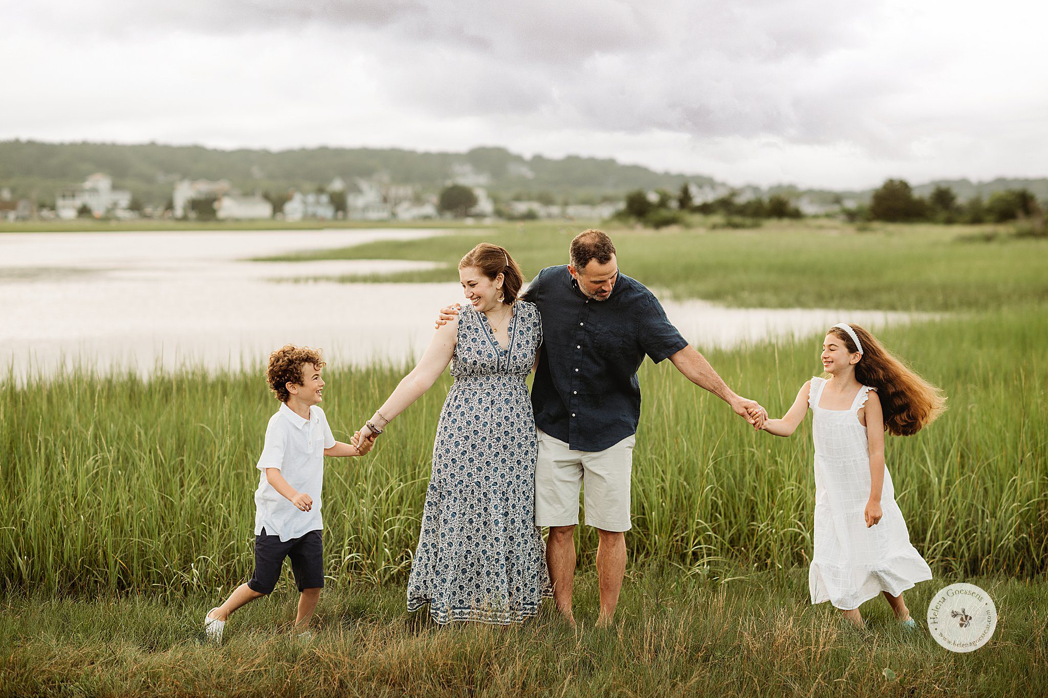 parents hug while kids hold their hands and run in circle in grass during Marshfield family portraits on the beach 