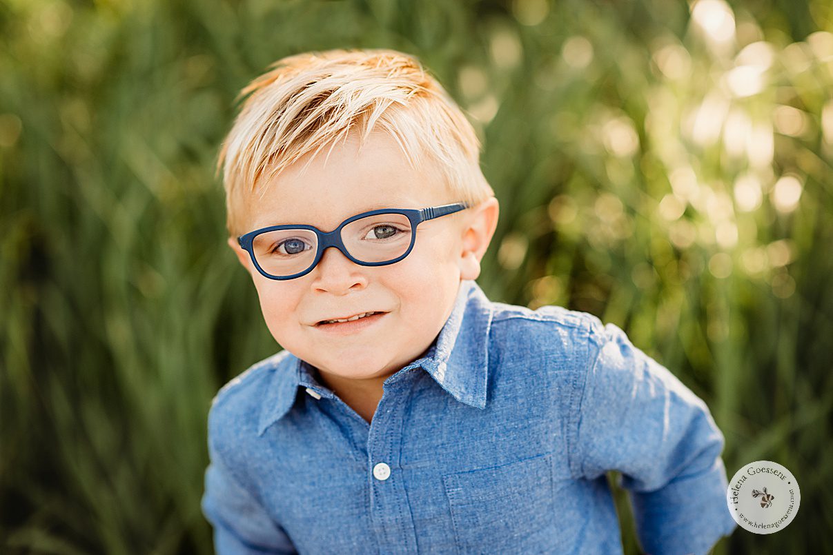 blonde boy in blue button up shirt with blue glasses smiles during family photos in Boston MA