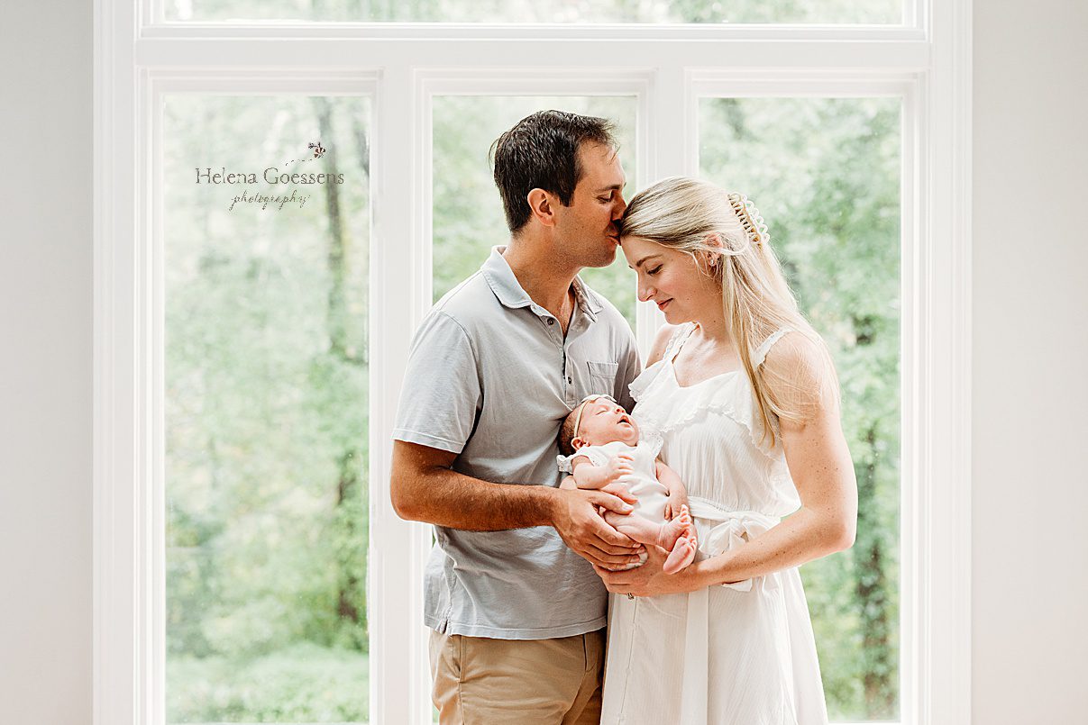 dad kisses mom's forehead while holding onto baby girl in white dress during lifestyle newborn photos in Westwood MA