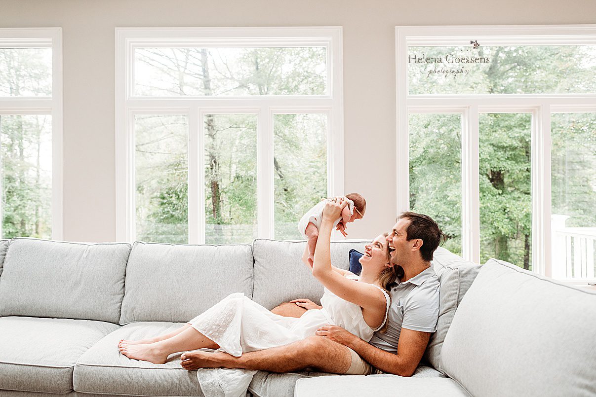 mom sits between dad's legs holding up baby girl on couch during lifestyle newborn photos in Westwood MA