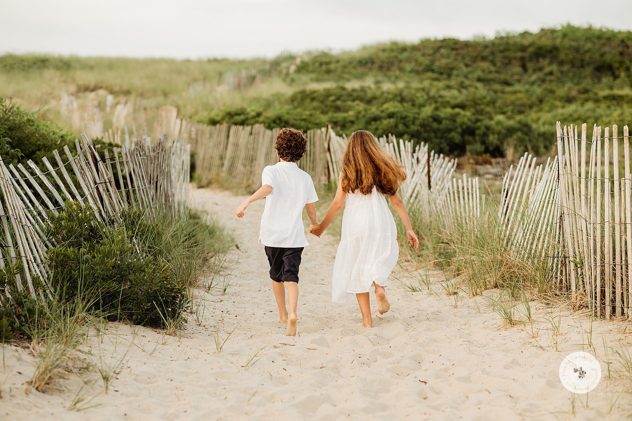 siblings hold hands running through sand dunes and wooden fences during Marshfield family portraits on the beach 