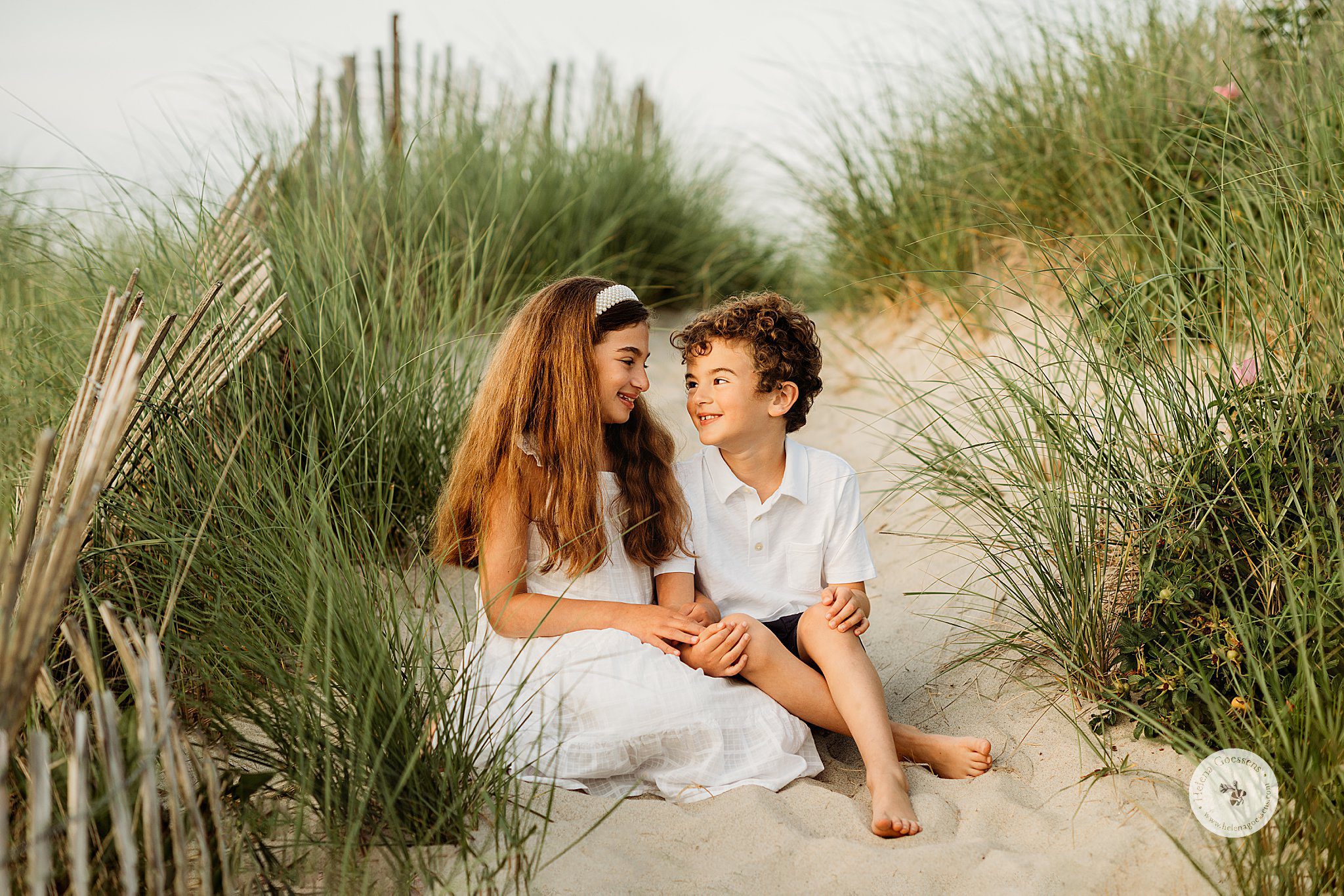 siblings in white outfits sit on sand dunes between sea grass during Marshfield family portraits on the beach 