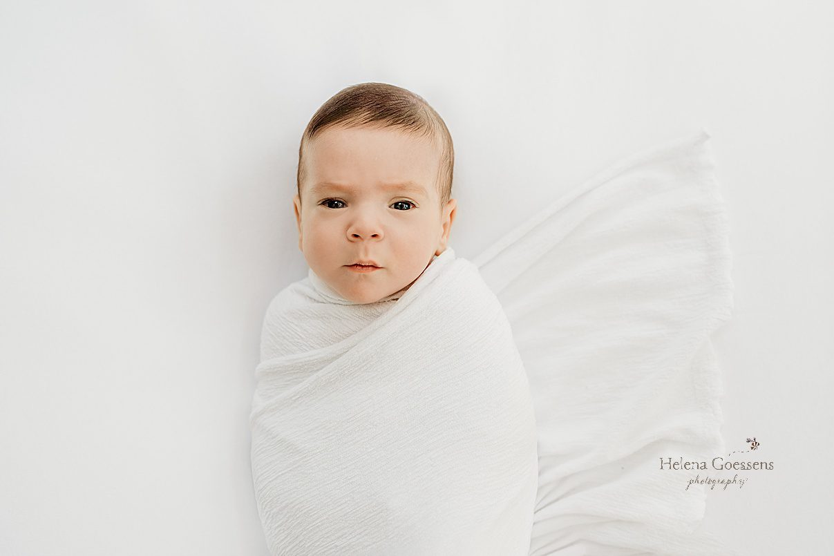 baby lays on bed in white wrap looking at camera during newborn photos at home in Boston MA