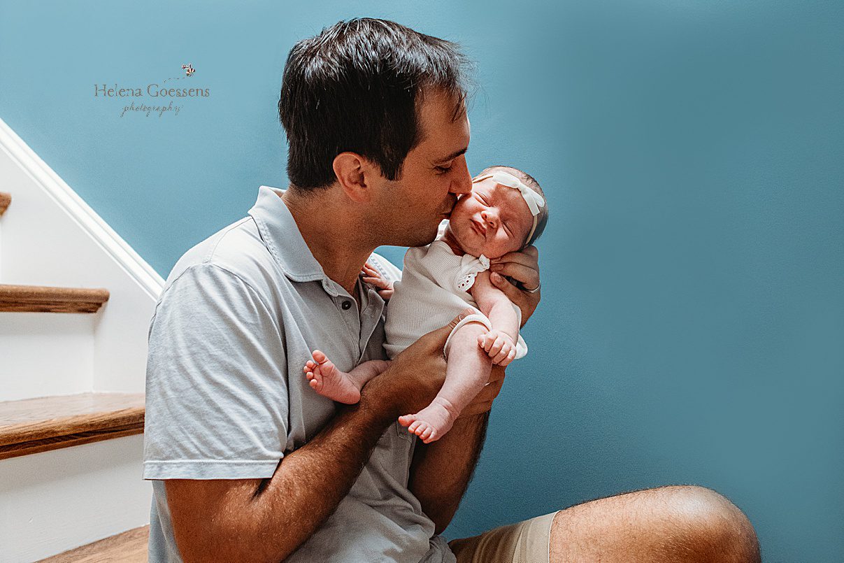dad kisses newborn daughter's cheek in front of blue wall during lifestyle newborn photos in Westwood MA