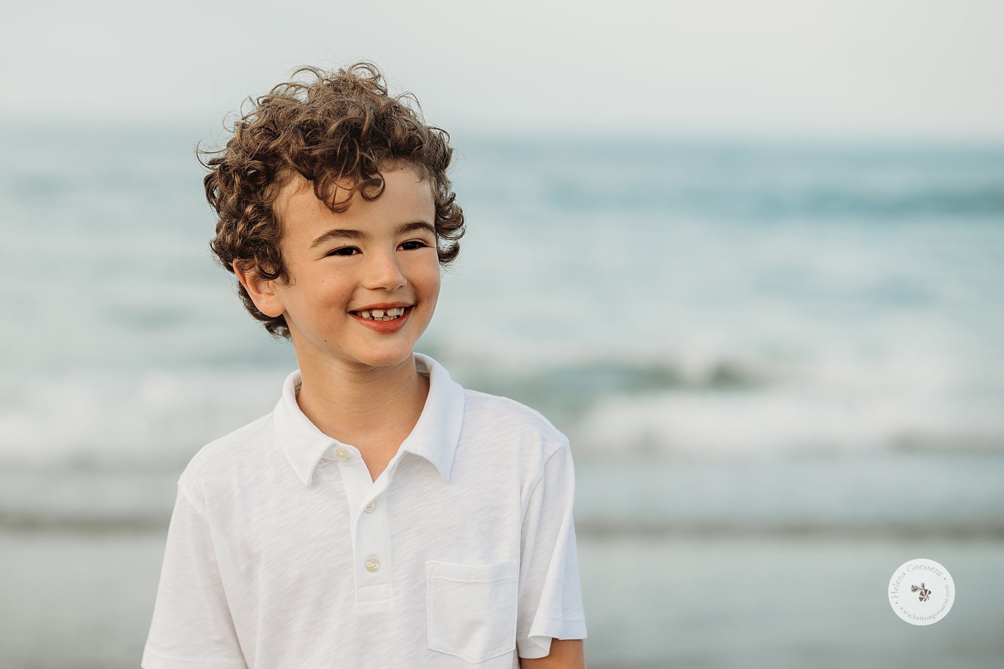boy with curly brown hair smiles in white polo shirt in front of water in Massachusetts 