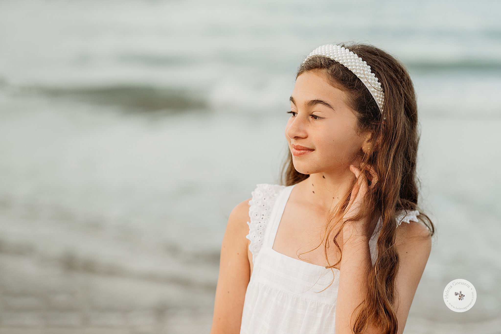girl in white dress with white pearl hand band pushes hair behind ear on beach during family photos 