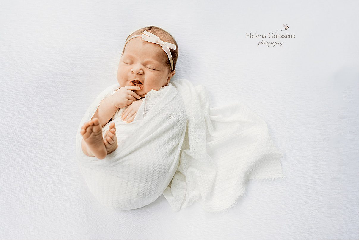 baby girl lays curled up in white wrap with small white headband during newborn photos at home 