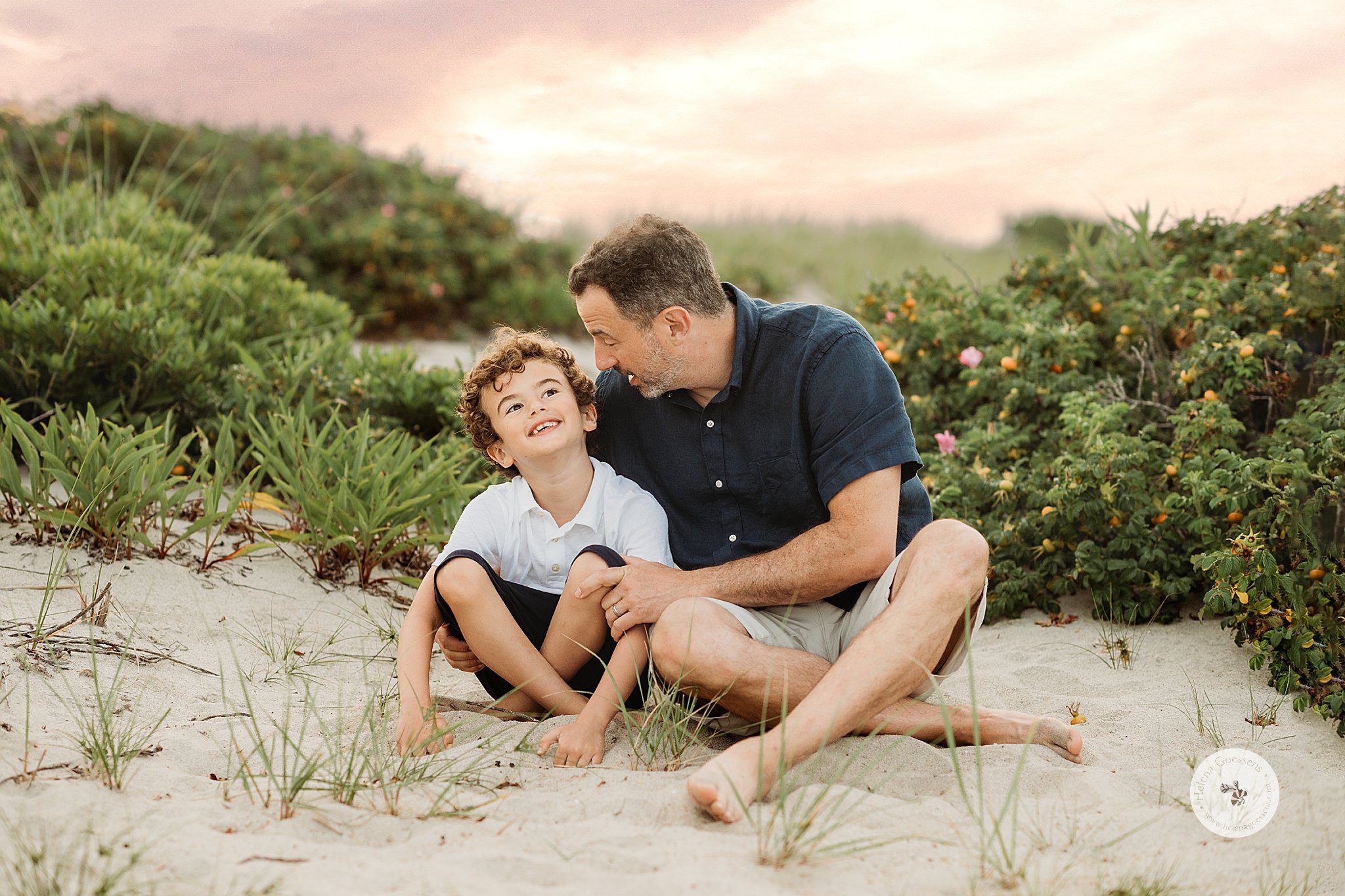 dad and son sit on sand dune with pink and yellow sunset during Marshfield family portraits on the beach 