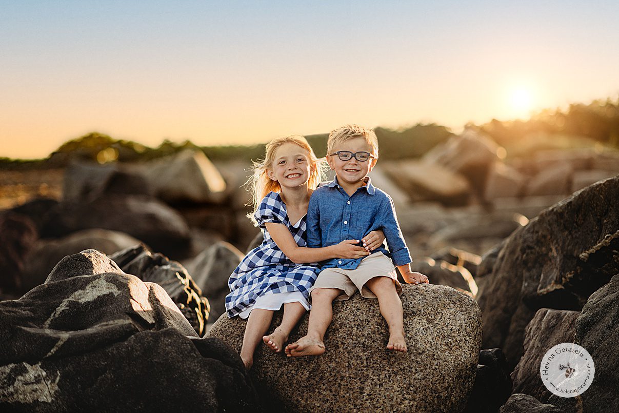 blonde children in blue and white outfits sit on rocks hugging during beach family portraits in Swampscott MA