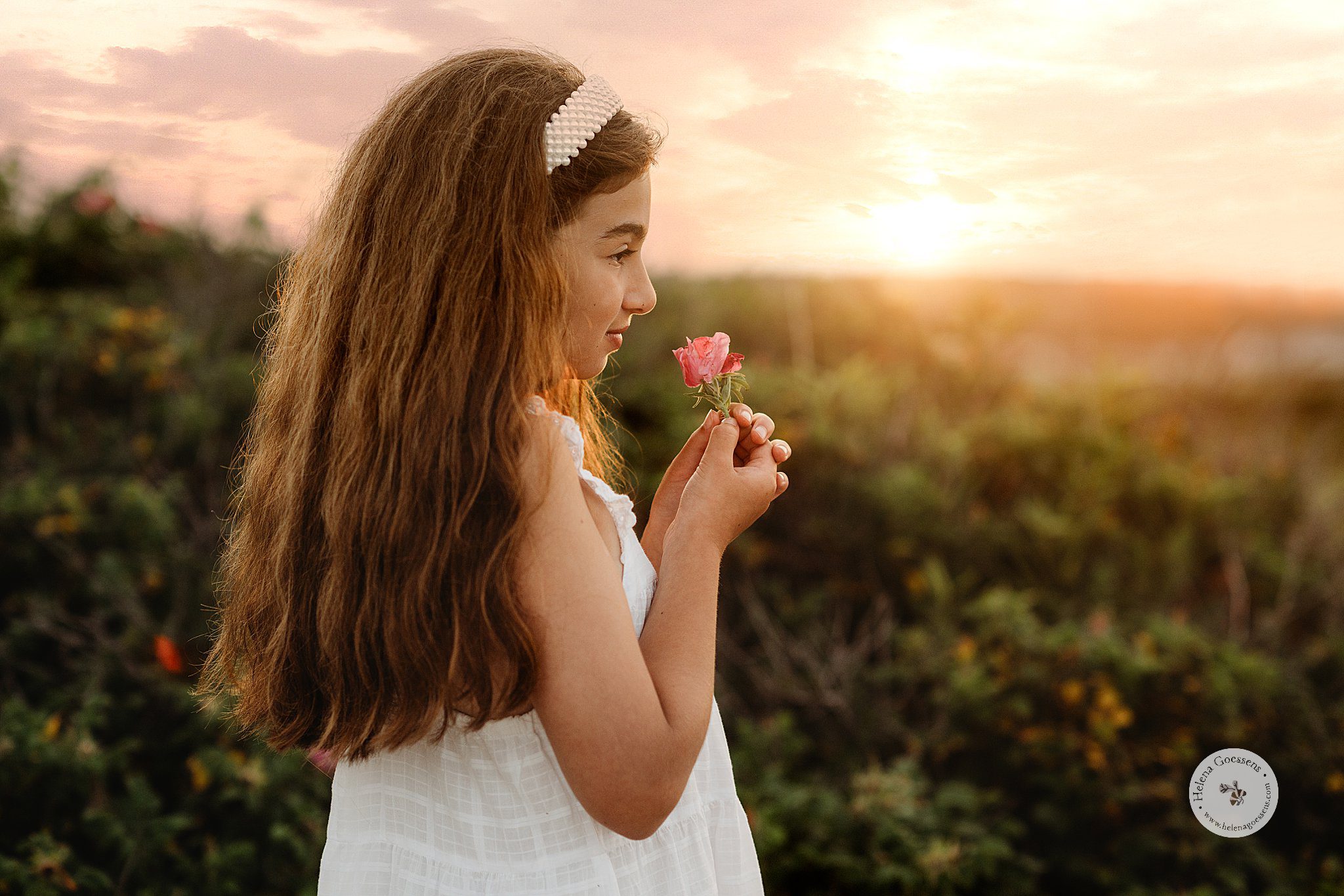 girl with long brown hair smells pink flower in white dress on beach 