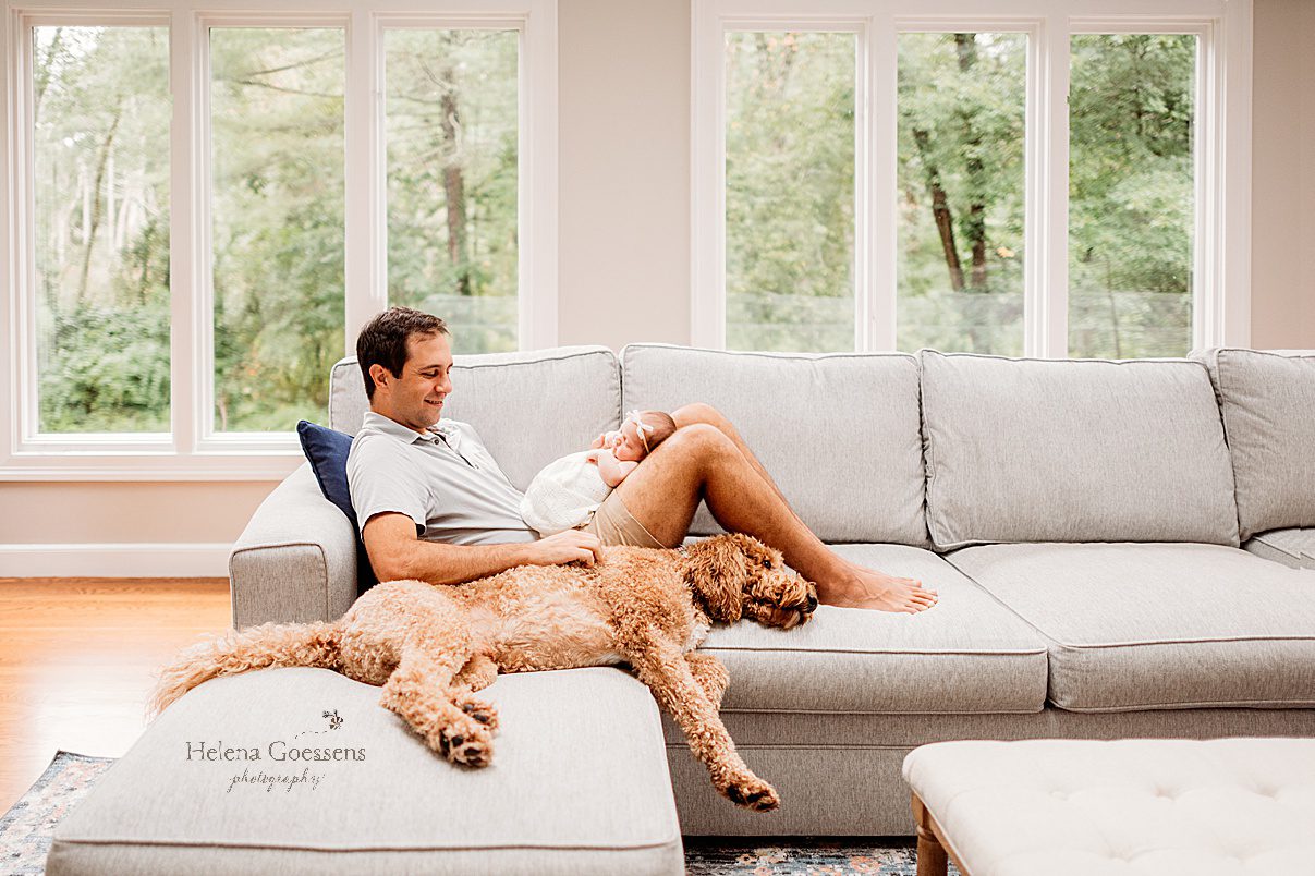 dad sits on couch with newborn daughter on his lap and dog sleeping next to him during lifestyle photos at home 