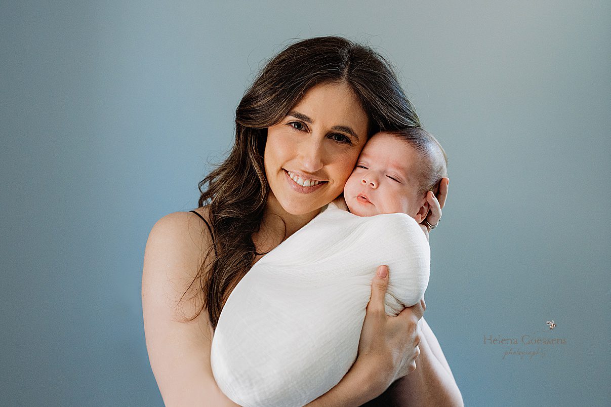 brown haired woman holds baby boy swaddled in white wrap in front of blue wall 
