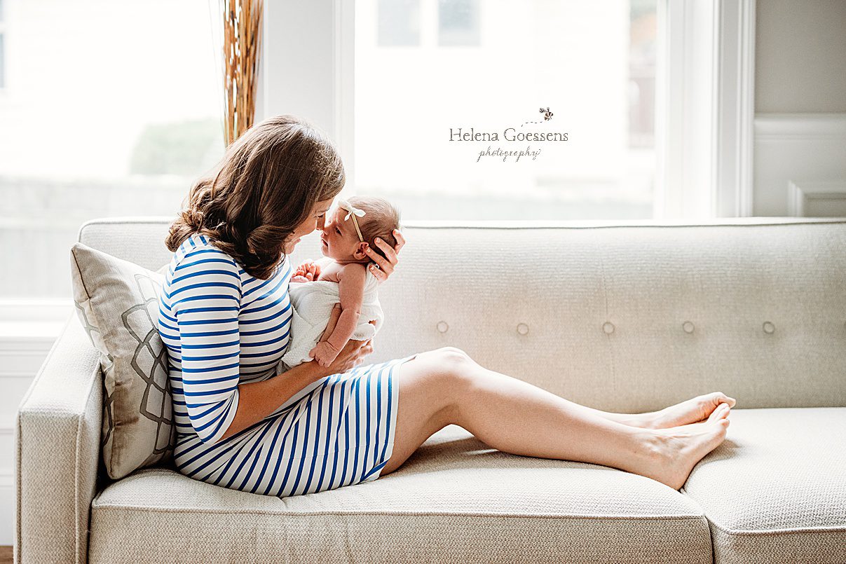 mom in blue and white striped dress sits on couch holding newborn baby girl nuzzling her nose