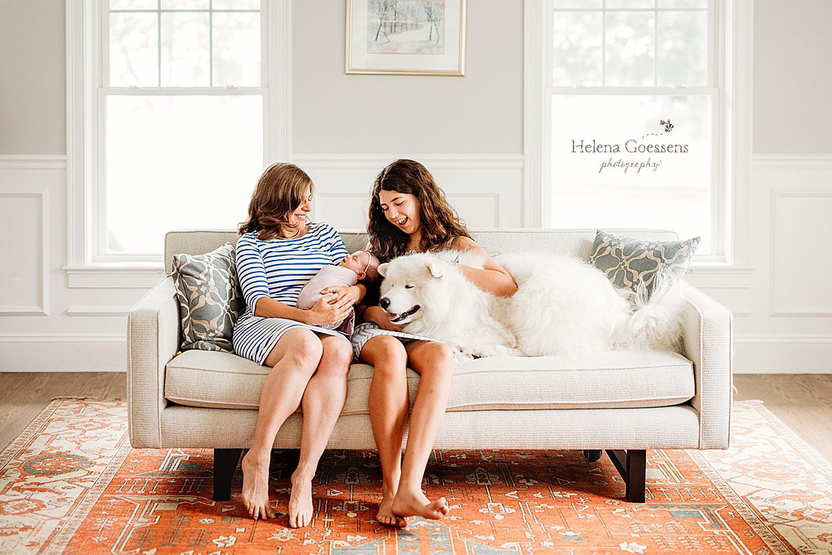 mom sits on couch with oldest daughter and newborn baby girl white dog during Needham MA lifestyle newborn portraits