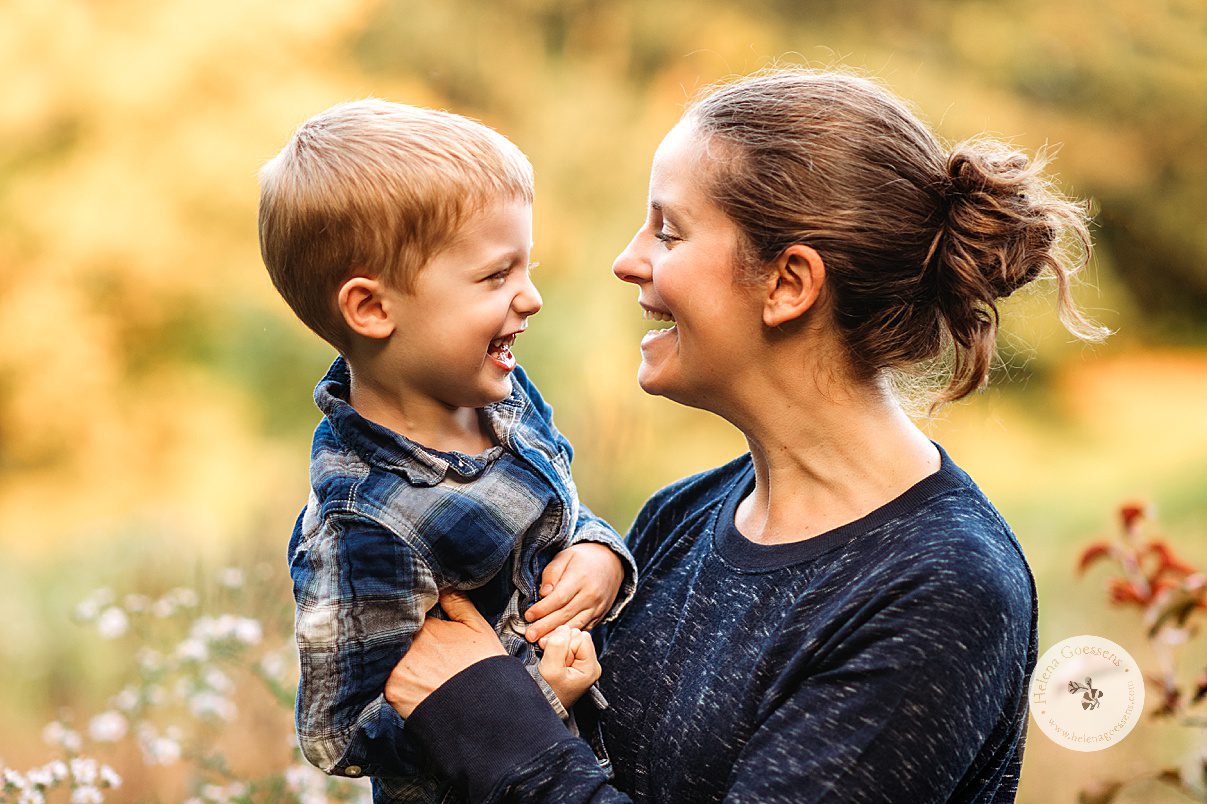 mom stands holding toddler in blue plaid shirt during fall Larz Anderson Park family portraits