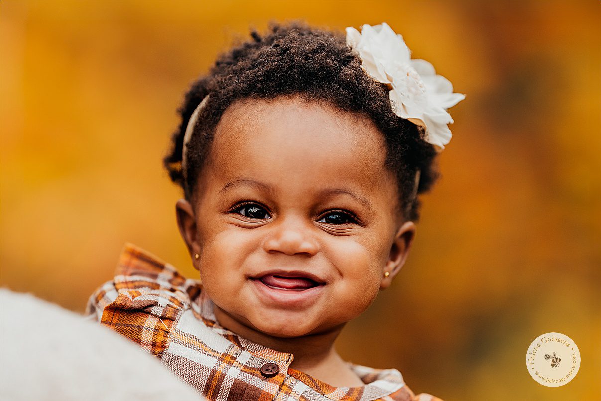 black toddler smiles with white flower in hair during fall lifestyle family portraits in Westwood MA