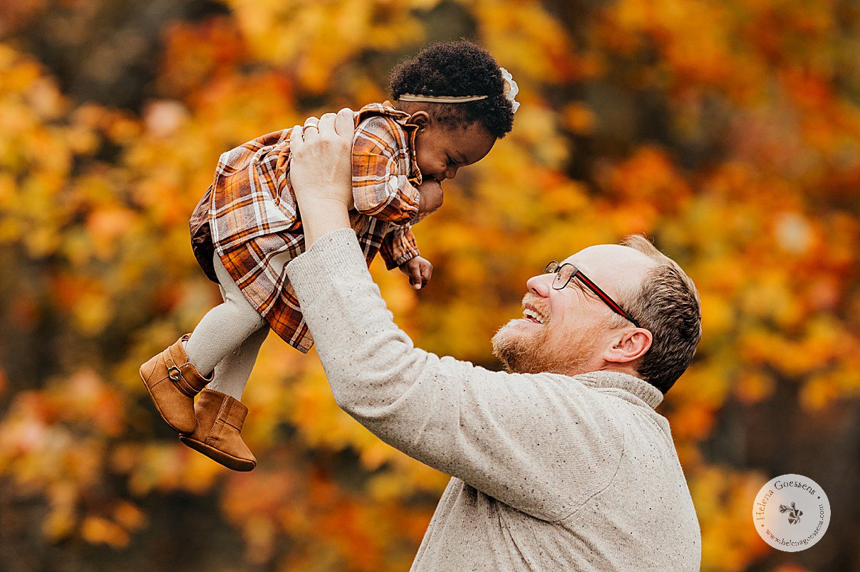 dad lifts up toddler in plaid dress in front of orange and yellow tress during fall lifestyle family portraits in Westwood MA