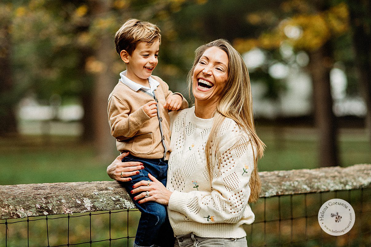 mom holds son on wooden fence during fall family portraits at the Bradley Estate while they laugh