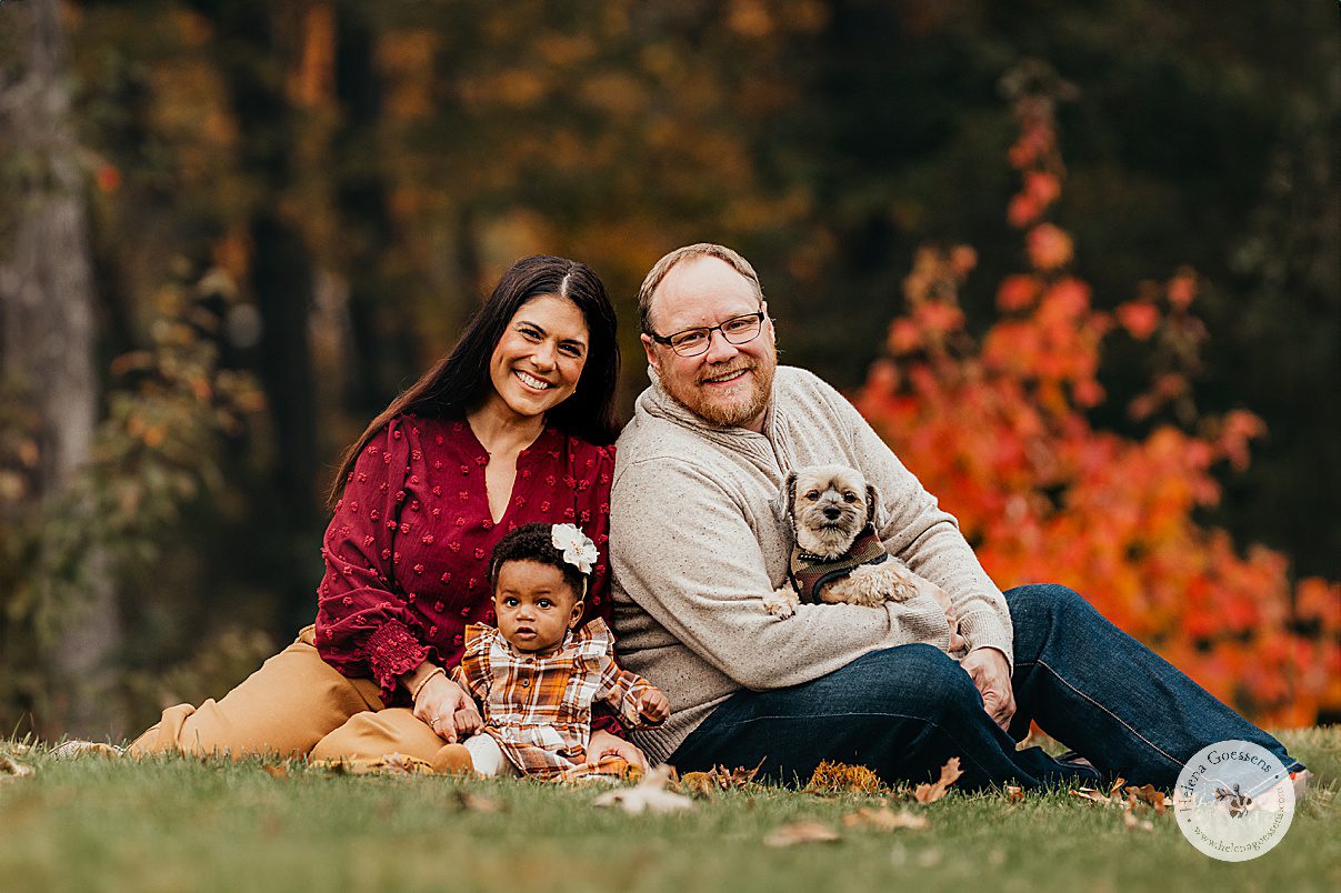 parents sit with black toddler between them in plaid fall dress while dad holds dog during fall lifestyle family portraits in Westwood MA