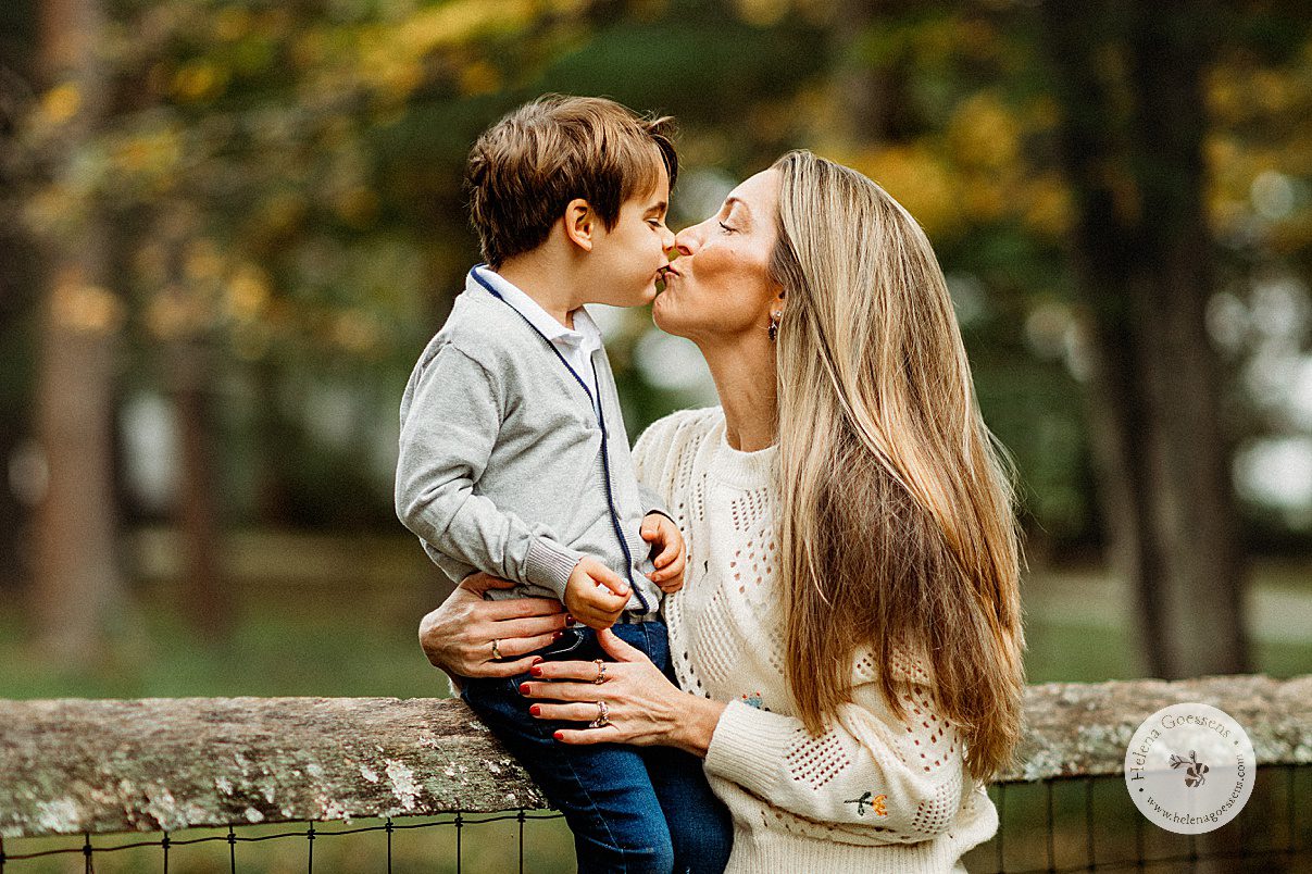 mom in ivory sweater leans to kiss toddler sitting on top of wooden fence during fall family portraits at the Bradley Estate