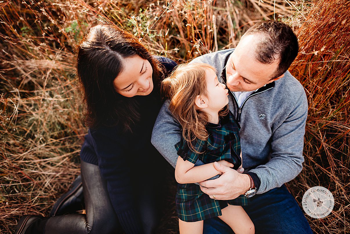parents sit in tall grass holding toddler in plaid dress during fall family portraits in Concord, MA